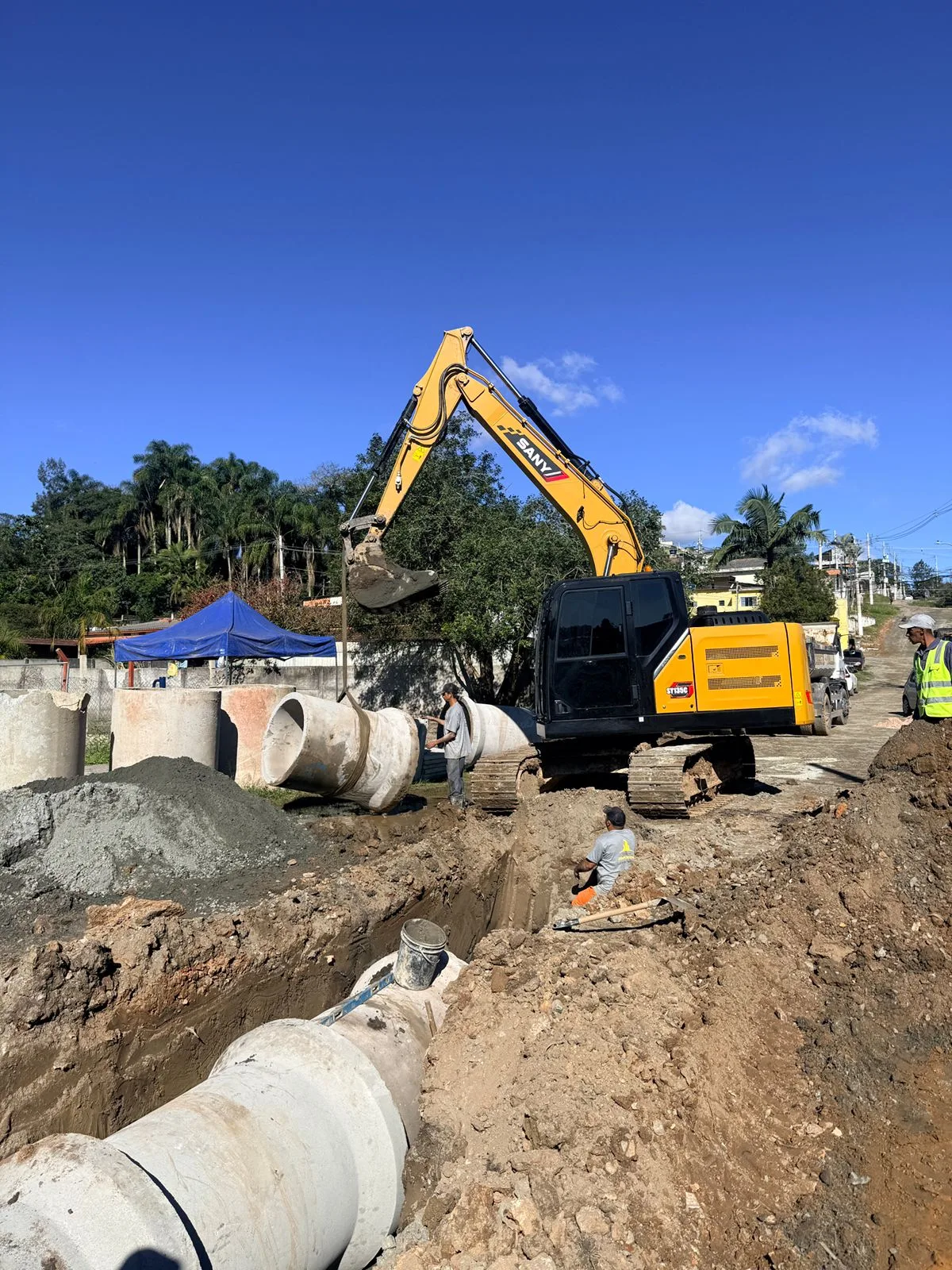 Escavadeira SANY assentando tubos de concreto de rede de água pluvial em vala aberta